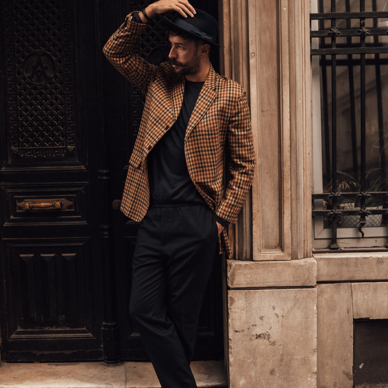 Man in a checkered jacket and black pants standing against a textured wall.